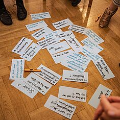 Mehrere Karteikarten mit handschriftlichen Notizen liegen auf dem Boden verteilt; darum ein Stuhlkreis