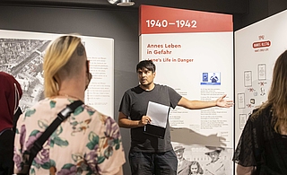 Ein Mann erklärt vor einer Ausstellungswand mit historischen Informationen einer kleinen Gruppe etwas und gestikuliert dabei.