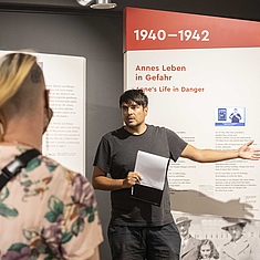 Ein Mann erklärt vor einer Ausstellungswand mit historischen Informationen einer kleinen Gruppe etwas und gestikuliert dabei.