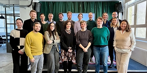 Eine Gruppe von 17 Personen, in zwei Reihen vor einer Bühne und der Kamera zugewandt für ein Gruppenfoto aufgestellt.