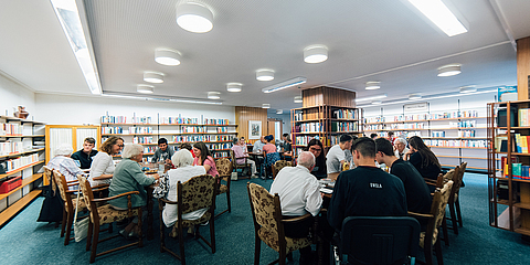 Ein Bibliotheksraum mit mehreren Gruppen von Menschen unterschiedlichen Alters, die an Tischen sitzen und sich unterhalten.